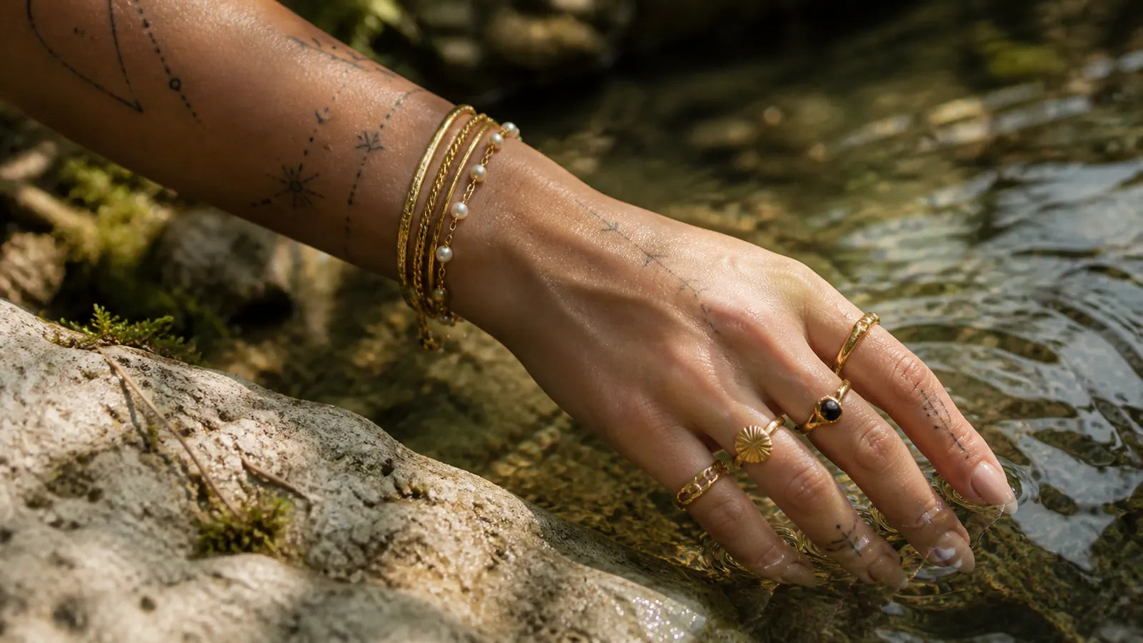 Tattooed hand wearing layered Arley London gold jewellery beside a woodland stream